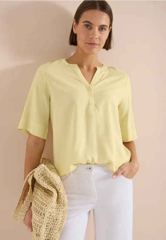 Frau in pastellgelber V-Bluse mit halblangen Ärmeln, weißer Hose und geflochtener Strohtasche vor hellem Hintergrund