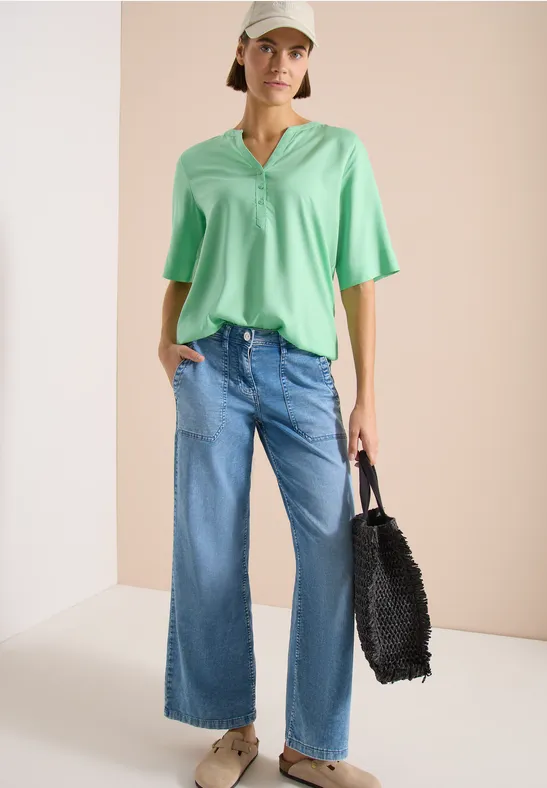Model in mintgrüner, lockerer V-Ausschnitt-Bluse, weiten Jeans und schwarzer Felltasche vor beigem Hintergrund.