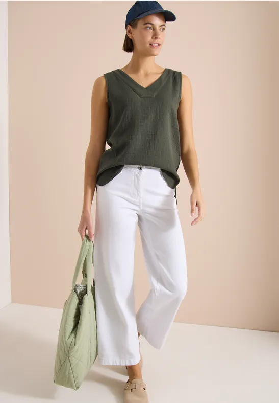 Person trägt weiße Culotte, olivgrünes ärmelloses V-Shirt, beige Sandalen und grüne Tasche.