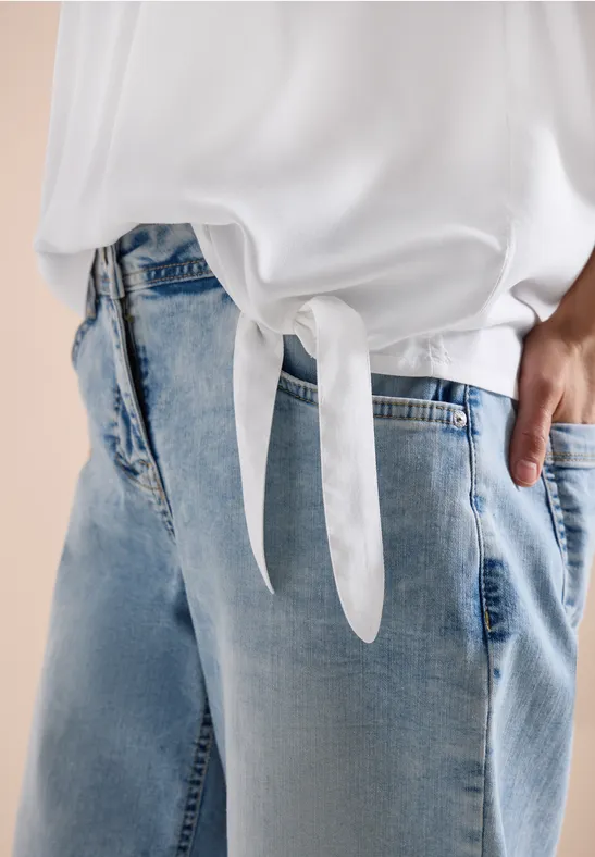 Person trägt weiße, vorne geknotete Bluse und helle Jeans, Hand in Tasche.