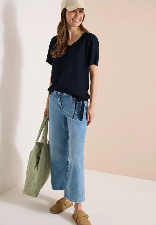 Vrouw met zwarte t-shirt, wijde spijkerbroek, bruine mules, en een grote groene stoffen tas in haar hand.