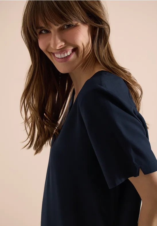 Zijaanzicht van een vrouw in donkerblau kort mouw top voor een lichte achtergrond.