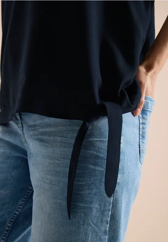 Persoon draagt blauwe jeans en donker top met zijstrik, hand in broekzak.