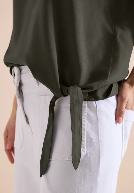 Eine Person in hellen Jeans trägt ein olivgrünes Oberteil, das seitlich geknotet ist.