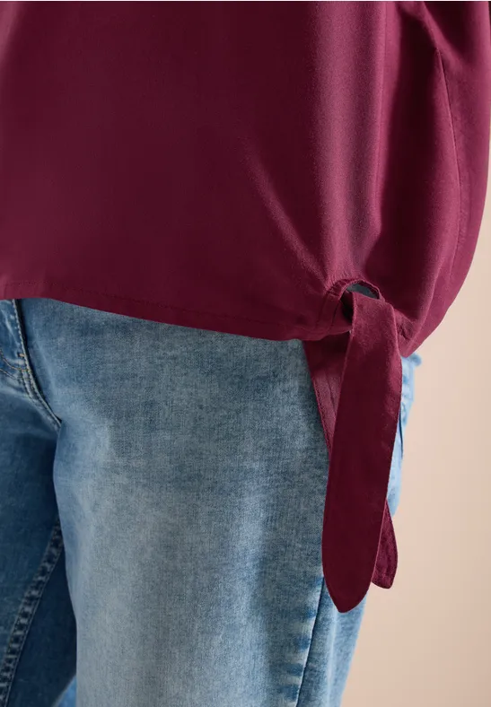 Hüftnahe Aufnahme: Jeans und ein seitlich geknotetes, burgunderrotes Oberteil.