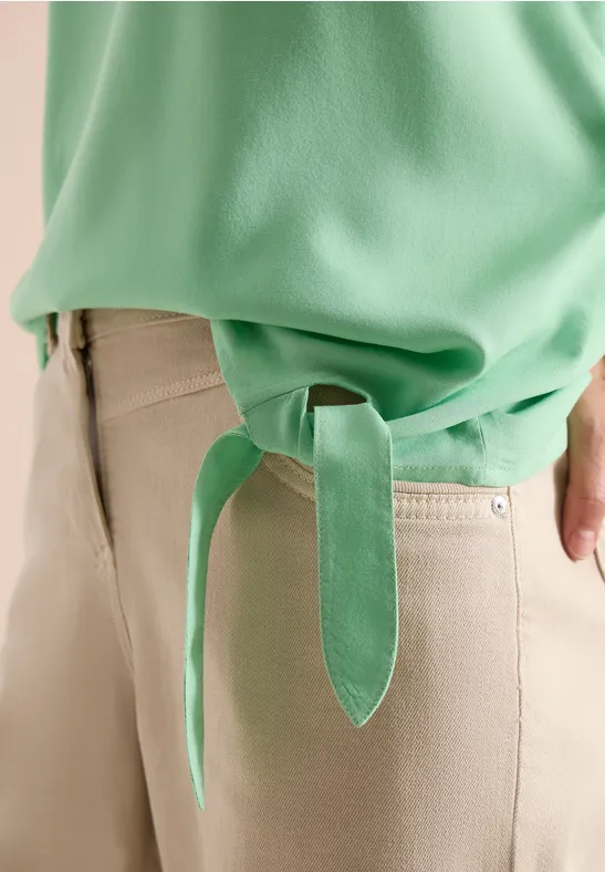 Nahaufnahme eines grünen Oberteils mit seitlicher Schlaufe und beiger Hose.