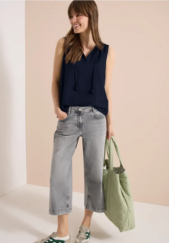 Frau in ärmelloser dunkelblauer Bluse, weiten grauen Jeans und grüner Stofftasche vor beigem Hintergrund.