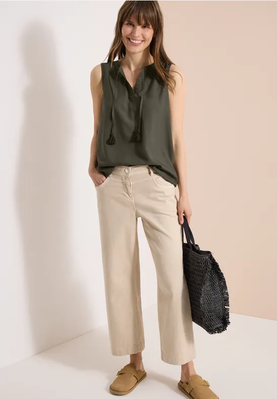 Frau trägt olivgrünes ärmelloses Top, helle weite Cropped-Hose, cognacfarbene Loafers und schwarzen Korb-Tote.