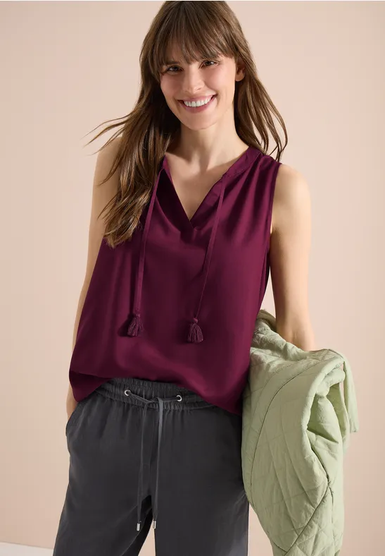 Eine Frau in einem ärmellosen, weinroten Oberteil und grauer Hose hält eine grüne Jacke