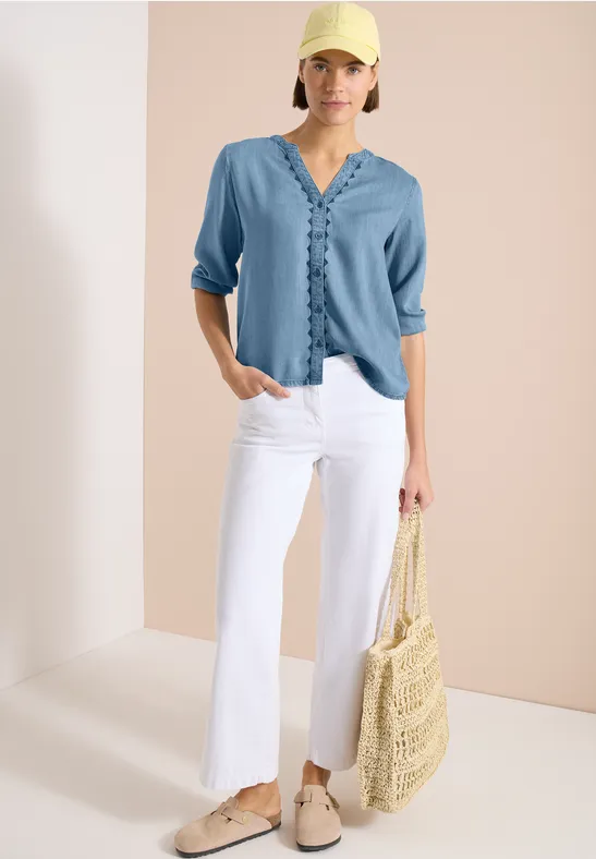 Model trägt hellblaue Bluse, weiße Schlaghose, beigefarbene Schuhe und hält eine Strohtasche.
