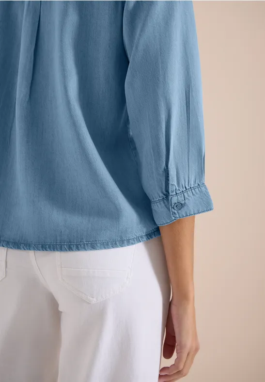 Seitliche Rückansicht einer Person in hellblauer, 3/4-Arm-Bluse und weißer Hose.