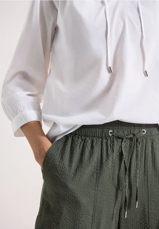 Weiße Bluse mit 3/4-Ärmeln und dunkelgrüner, strukturierter Hose mit Tunnelzug und Taschen.