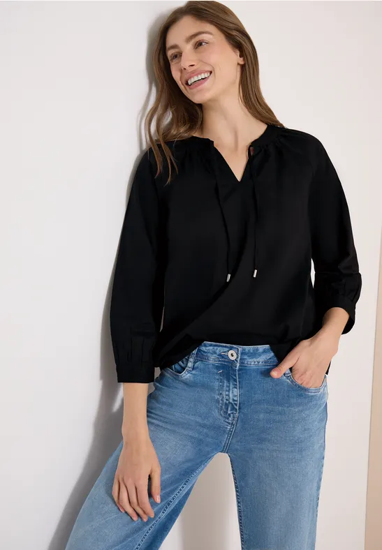 Frau in schwarzem Pullover mit Kapuze und blauen Jeans lehnt sich lässig an weiße Wand.