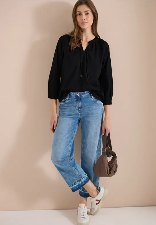 Person in schwarzer Bluse, blauen Jeans und weißen Sneakern steht mit grauer Tasche vor beige Wand.