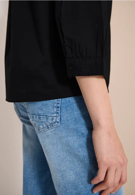 Eine Person trägt eine schwarze Bluse mit 3/4-Ärmeln und hellblaue Jeans, seitliche Nahaufnahme.