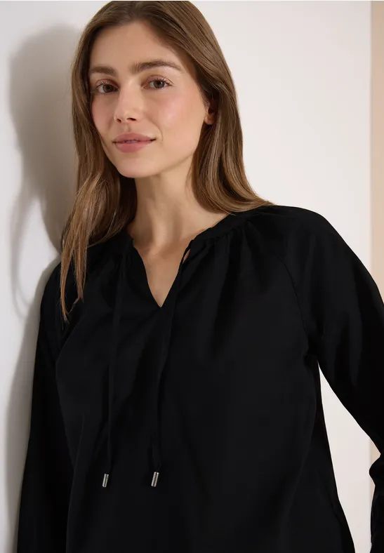 Eine Frau in einer schwarzen Bluse lehnt lässig an der Wand.