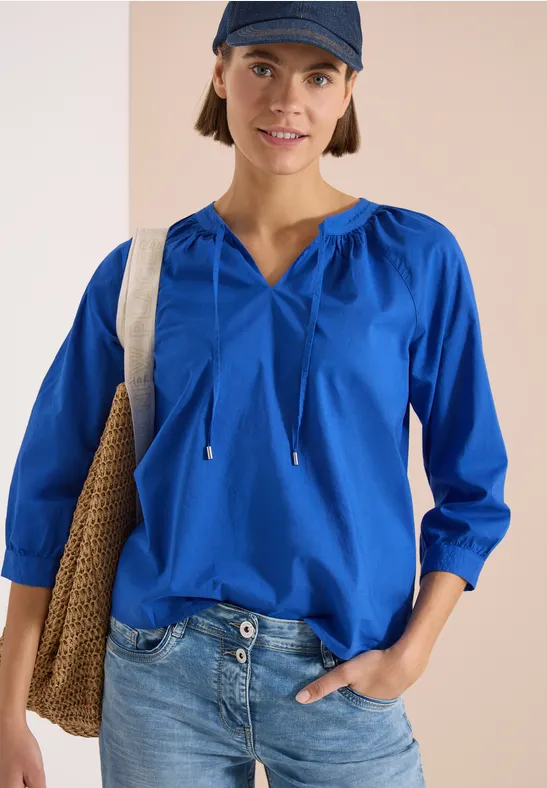 Person trägt blaue Bluse mit Kordeln, Jeans und Tasche vor neutralem Hintergrund.