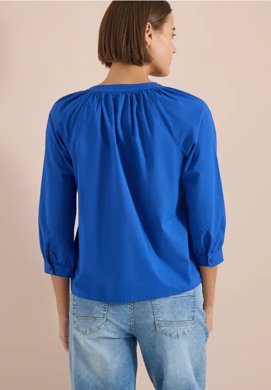 Frau von hinten in blauer, geraffter 3/4-Arm-Bluse und heller Jeans.