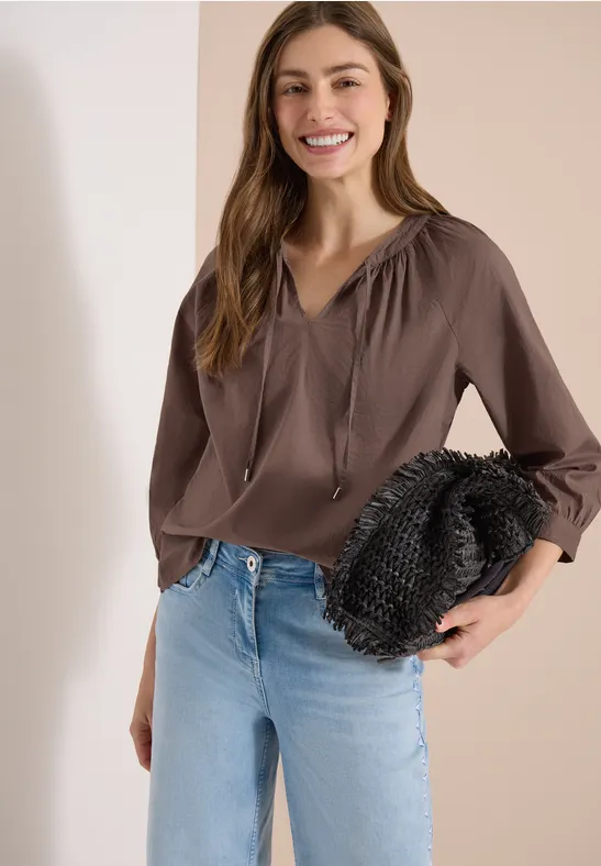 Frau mit brauner Bluse und Jeans hält schwarze Clutch vor hellem Hintergrund.