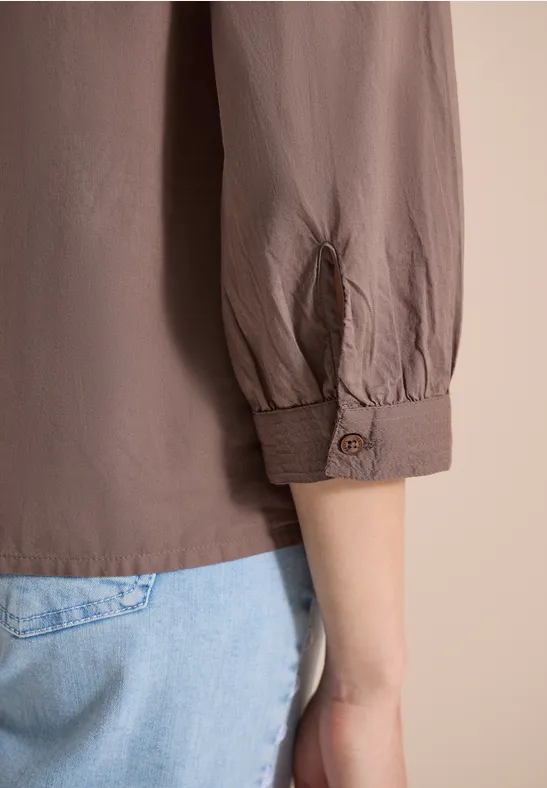 Ein Arm in taupefarbener Bluse mit hochgekrempeltem Ärmel und hellblauer Jeans vor hellem Hintergrund.
