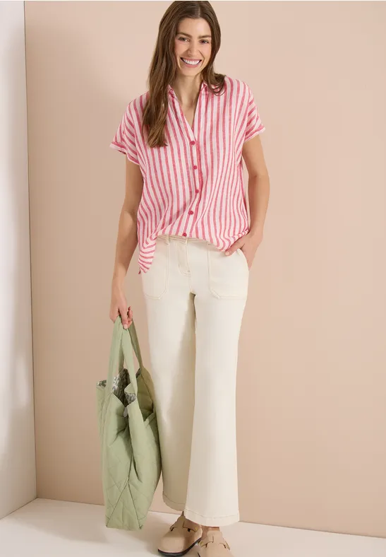 Frau trägt rot-weiß gestreiftes Hemd, weiße Hose und hält eine grüne Tasche vor beiger Wand.