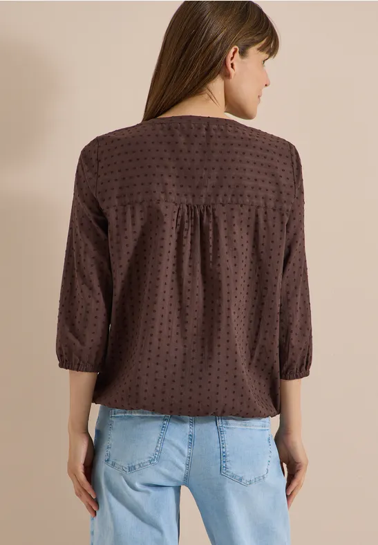 Rückenansicht einer Frau in gepunkteter brauner Bluse mit 3/4-Ärmeln und hellblauen Jeans.