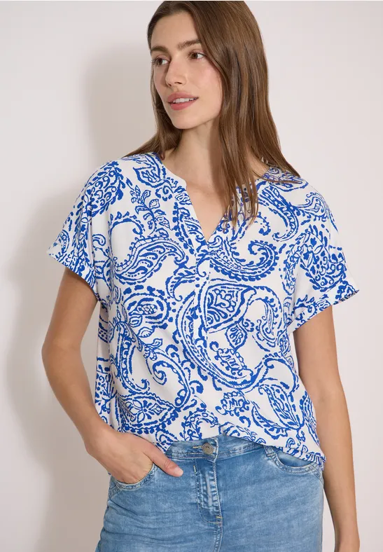 Vrouw draagt wit T-shirt met blauw paisleypatroon en spijkerbroek, hand in broekzak, neutrale achtergrond.