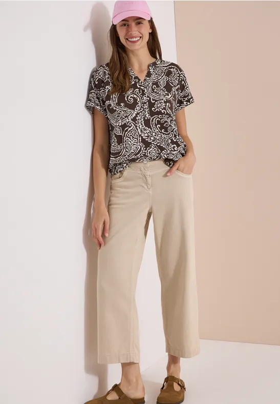 Vrouw leunt tegen tweekleurige achtergrond, draagt een blouse met print, wijde beige broek en slingback-pumps.