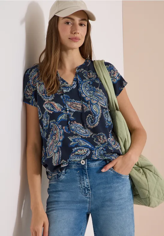 Vrouw leunend tegen een muur in een blauw-groen paisley blouse, spijkerbroek en groen gewatteerd tas.