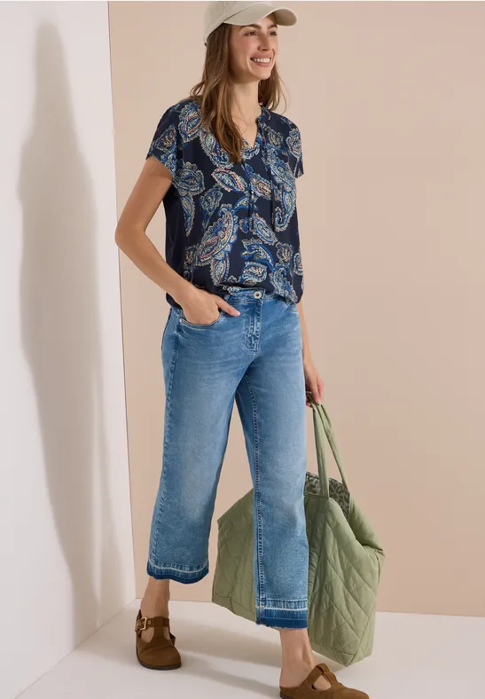 Vrouw met beige pet, gekleurde korte blouse, wijd korte spijkerbroek en grote groene gewatteerde tas.