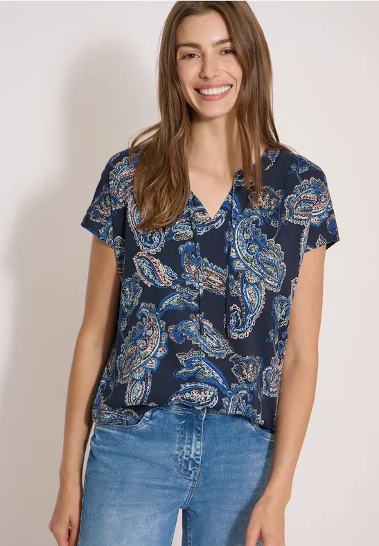 Jonge vrouw voor neutrale achtergrond, draagt een blauw shirt met paisley patroon en blauwe jeans.