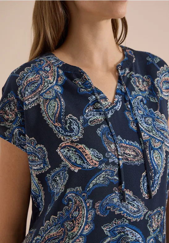 Damesblouse met korte mouwen en blauw-oranje paisleyprint op een donkere ondergrond.