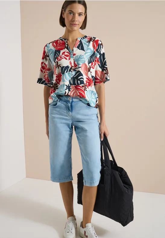 Person trägt ein buntes Hemd mit Blumenmuster, knielange Jeans-Shorts und hält eine große schwarze Tasche.