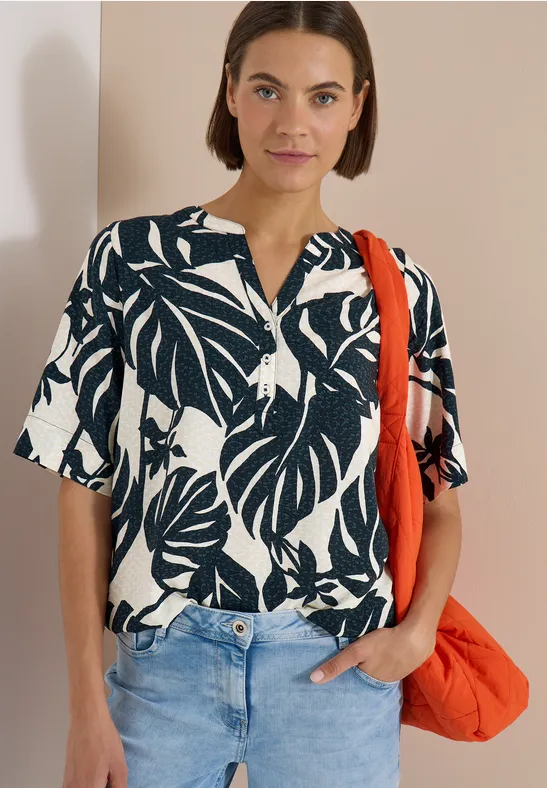 Frau mit schwarz-weißem Blusen-Shirt mit Blattmuster, Jeans und orangefarbener Tasche vor beigem Hintergrund.