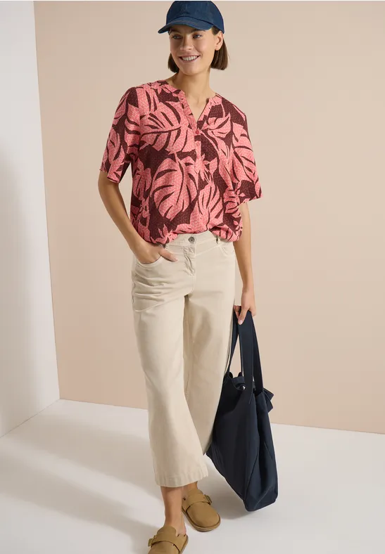 Model trägt rosa-schwarze Bluse mit Blattmuster, helle Culotte, beige Schuhe, blaue Tasche und Mütze.