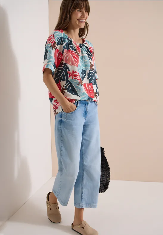 Frau in tropischer Print-Bluse, weiten Jeans, Plateauschuhen und schwarzer Tasche vor neutralem Hintergrund.
