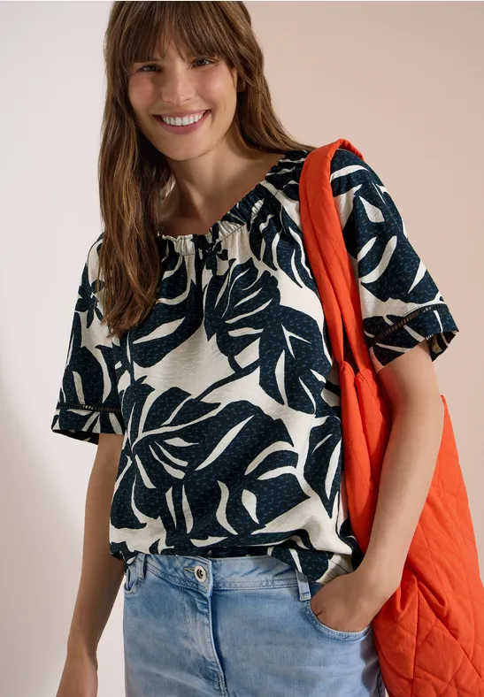 Vrouw draagt jeans en een wit T-shirt met donkerblauw bladpatroon, met een rode schoudertas.