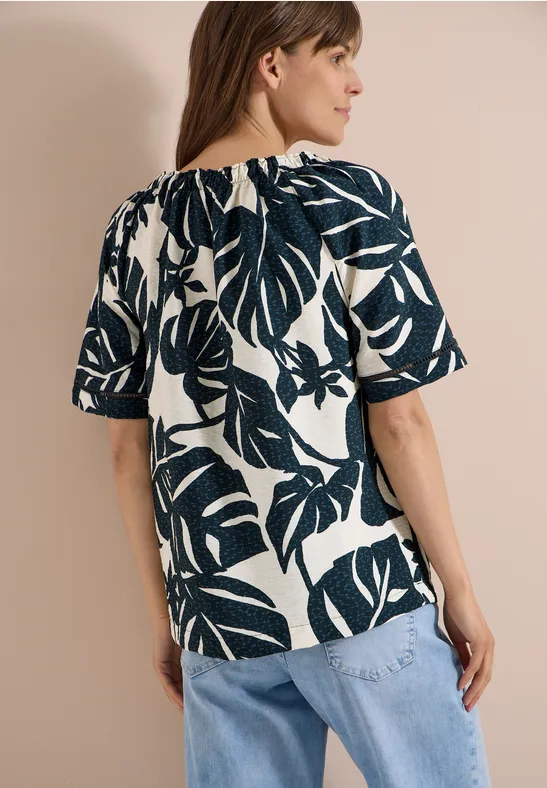 Vrouw van achteren in wit blouse met grote donkergroene bladeren en halflange mouwen, lichtblauwe jeans.