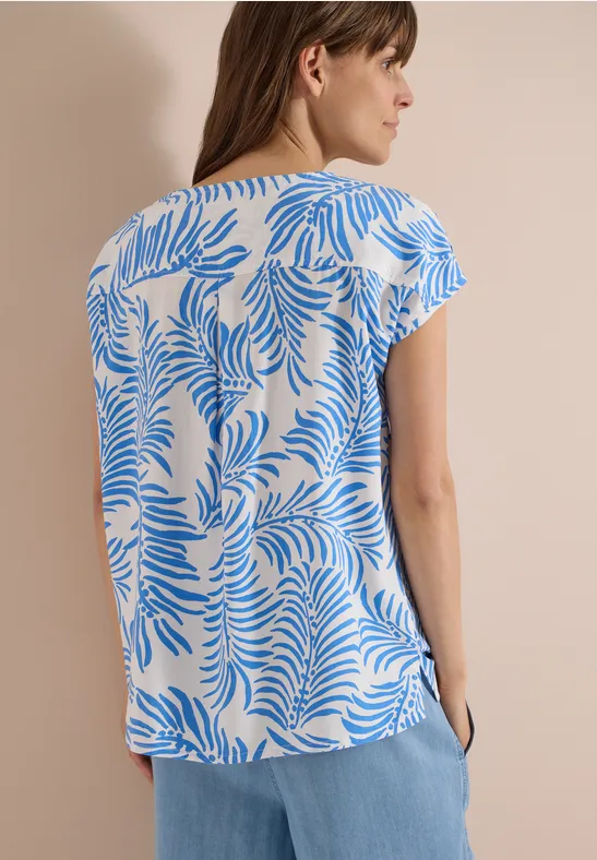 Vrouw van achteren in een wit-blauw dessinblouse en lichtblauwe broek voor een neutrale achtergrond.