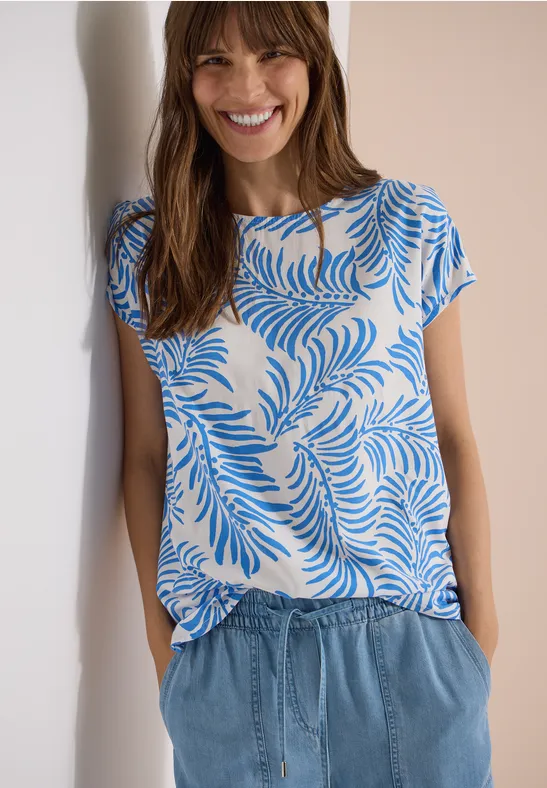 Vrouw in witte top met blauw bladerenpatroon en blauwe jeans, handen in de zakken.