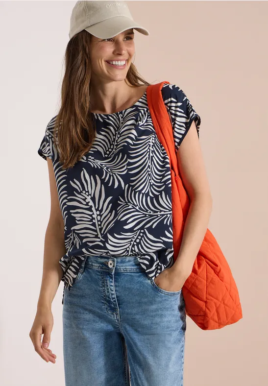 Vrouw in jeans, marineblauw bladprint-shirt en pet met een oranje gewatteerde tas.