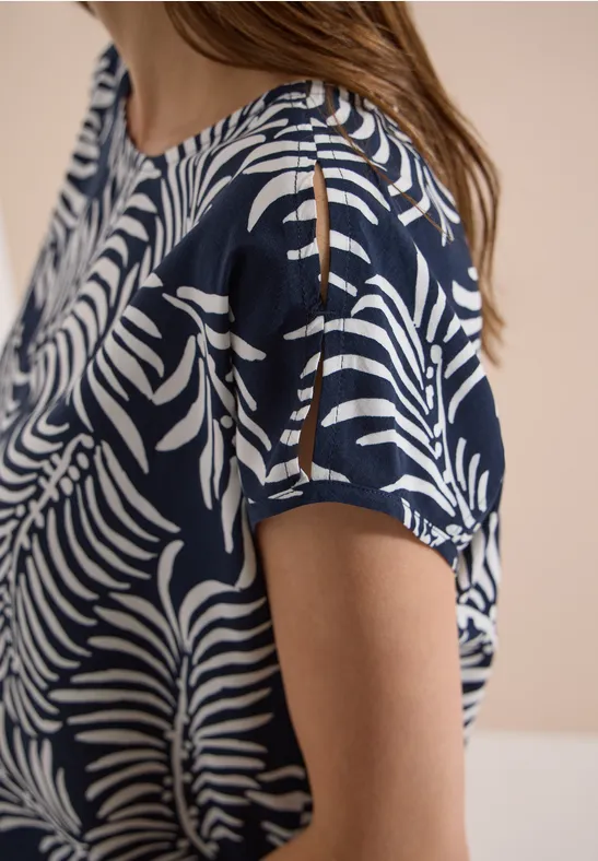 Vrouw draagt donkerblauw shirt met wit bladpatroon, korte mouwen en openingen op de schouders.