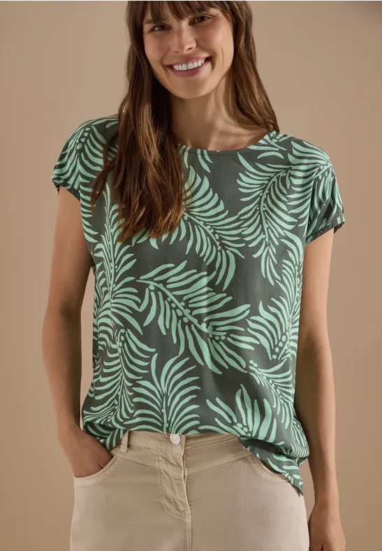 Een vrouw met wazig gezicht draagt een groen T-shirt met bladmotief en een lichte broek.