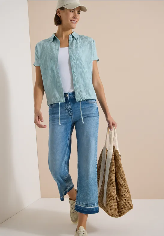 Frau in sommerlicher Freizeitkleidung: grünes Hemd, weißes Top, weite Jeans, Strohtasche und Sandalen.
