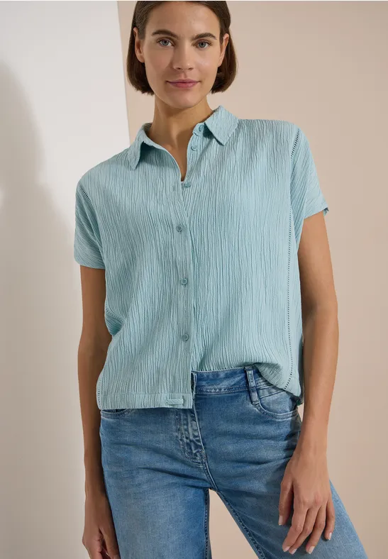 Eine Frau trägt eine hellblaue, kurzärmelige Bluse und Jeans vor einem ruhigen Hintergrund.