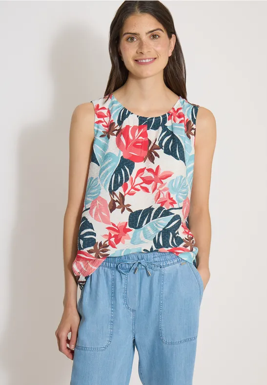 Vrouw draagt mouwloze, kleurrijke bloementop en blauwe broek, staat met handen in de zakken.