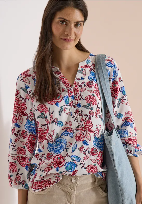 Frau trägt weiße Bluse mit roten und blauen Blumen, 3/4-Ärmel, dazu blaue Denim-Tasche.