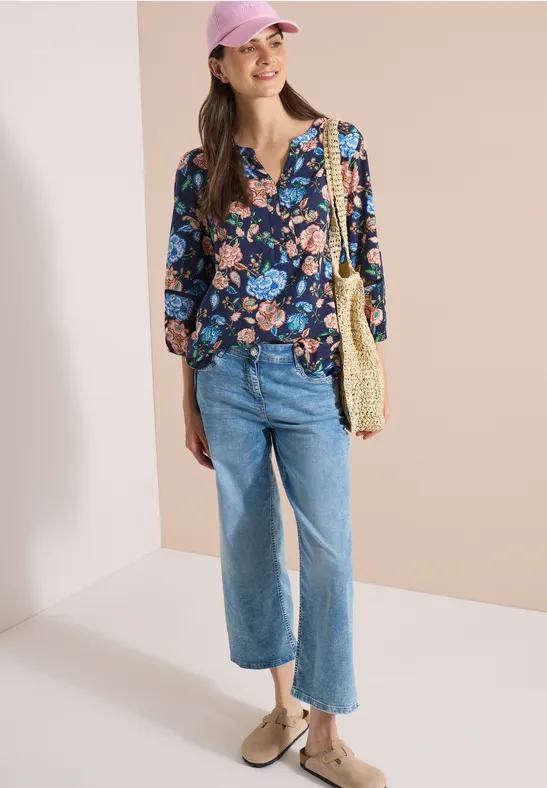 Eine Frau trägt eine lockere, blau florale Bluse, gerade Jeans, Basttasche und beige Loafers vor hellem Hintergrund.