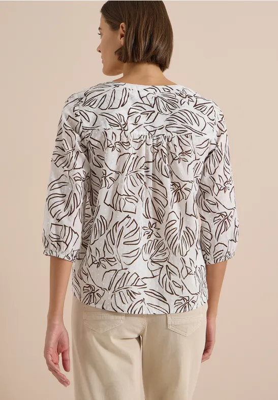 Frau von hinten in weißer Bluse mit schwarzem Blattmuster und hellen Hosen.