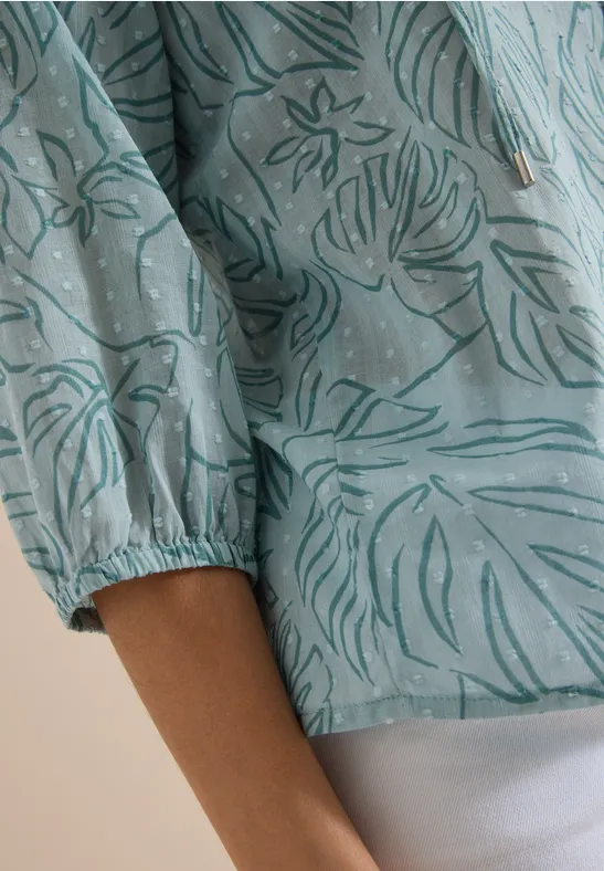 Nahaufnahme einer Person mit türkiser Bluse mit Blumenmuster und weißer Hose.